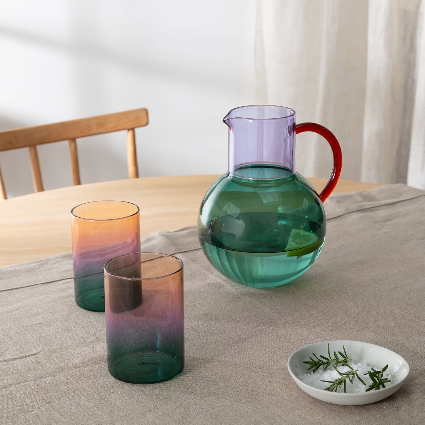 Colorful glass pitcher and tumblers on a table with a dish of herbs.