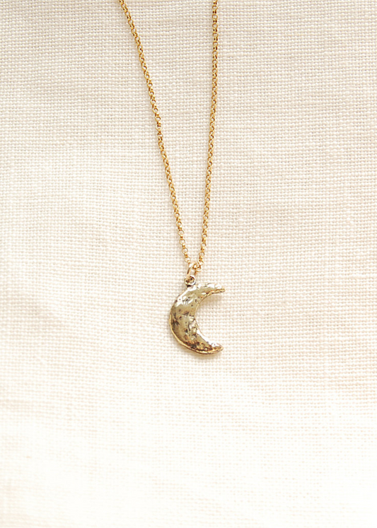 A gold-colored necklace with a hand-carved crescent moon pendant, displayed against a neutral background.