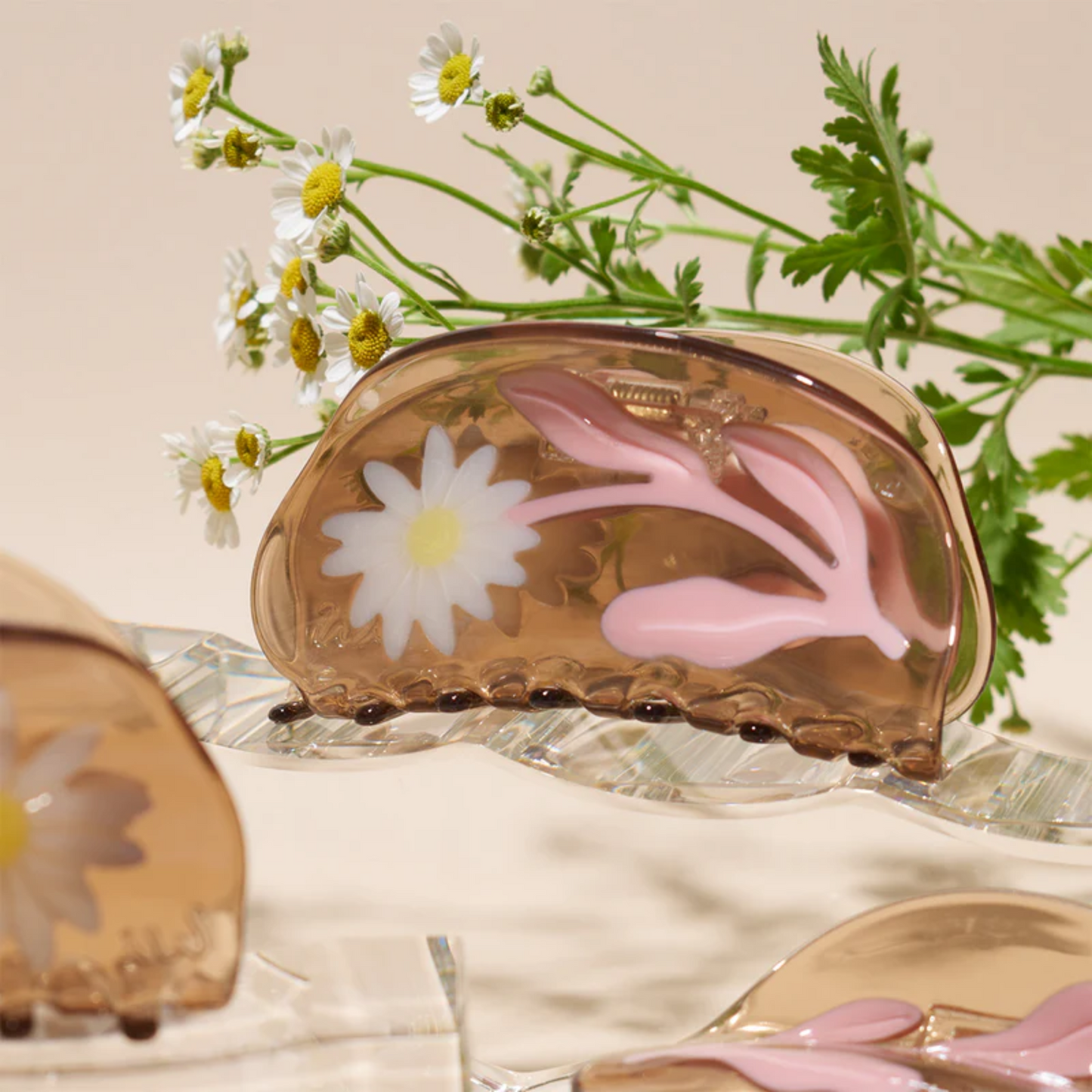 A beige hair claw with a floral pattern, featuring a prominent white flower and pink leaves, displayed against a neutral background with a few scattered real flowers.